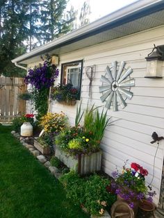 a house with lots of flowers and plants in the front yard, along with a clock on the side of the house