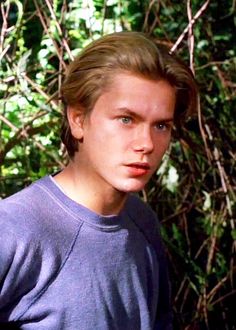 the young man is posing in front of some bushes and trees with his head turned to the side