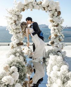 a bride and groom kissing in front of an arch of flowers with the ocean in the background