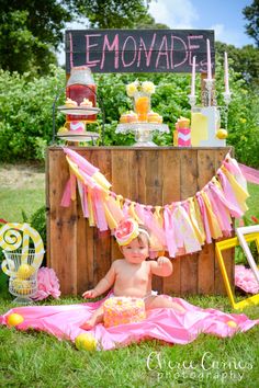 a baby sitting on the grass in front of a lemonade stand with pink and yellow decorations