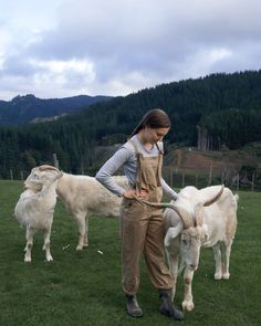 a woman in overalls standing next to two white cows on a lush green field