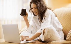 a woman sitting on a couch holding a coffee mug while using her laptop