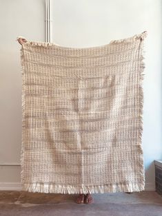 a woman standing in front of a white wall holding up a beige blanket with fringes