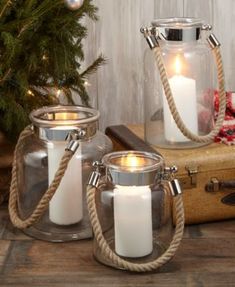 two candles are sitting in glass containers next to a christmas tree and suitcase on a wooden table