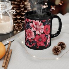 a black coffee mug with pink flowers on it next to some pine cones and candles