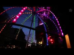 a ferris wheel lit up at night with colorful lights in the sky and buildings behind it