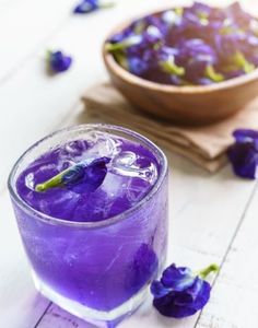 a purple drink in a glass next to a bowl of flowers