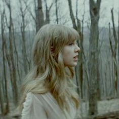 a woman with long blonde hair standing in the woods looking off into the distance,