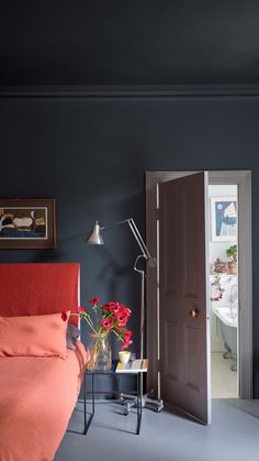 a bedroom with dark blue walls and red bedding in the foreground, an open door leading to another room