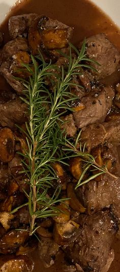 a bowl filled with meat and potatoes covered in gravy, garnished with a sprig of rosemary