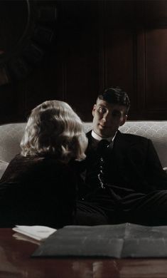 a man and woman sitting on a couch looking at each other in front of a large clock