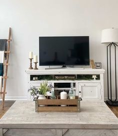 An engaging farmhouse living room is designed with the main focus on a charming wooden coffee table and a fabulous entertainment console. The aesthetic is amplified by a delicate blanket ladder draped with alternating gray and white throws. The table is garnished with a simple, crate-like centerpiece filled with glass bottles and greenery...   Image: mymidwestdiy