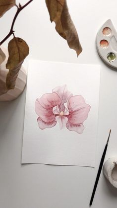 a watercolor painting of a pink flower on white paper next to paintbrushes