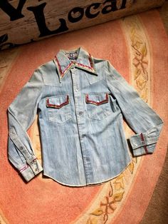 This is one amazing find! Light wash denim  snap shirt Rad Santa Fe Native American chiefs head image on back with sequin stitching on back on cuffs of sleeves and along collar and chest pockets Flap chest pockets with snap closures This is truly a unique piece There are several smaller rust stains on this shirt Through out the shirt Photos taken reveal the staining Tag reads size small Measurements are approximate and taken flat so please double bust and waist for more accurate sizing Bust: 16 Vintage Medium Wash Pre-washed Tops, Vintage Collared Tops For Rodeo, Vintage Medium Wash Tops For Rodeo, Vintage Denim Top For Rodeo, Retro Medium Wash Top For Fall, Vintage Denim Button-up Top, Vintage Medium Wash Top With Button Closure, Vintage Cotton Shirt For Festivals, Vintage Medium Wash Top With Buttons