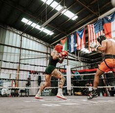 two men boxing in an indoor ring with flags hanging from the ceiling and one man wearing orange shorts