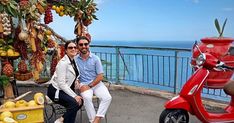 two people sitting on a bench next to a red scooter and fruit stand