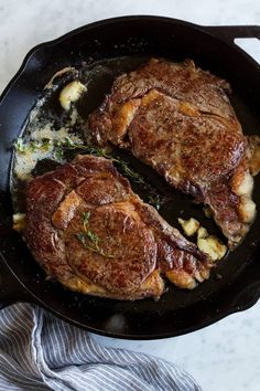 two steaks cooking in a skillet with potatoes and herbs on the side, ready to be cooked