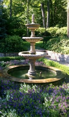 a water fountain surrounded by purple flowers and greenery in a park setting with trees