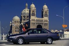 two people standing on the hood of a car in front of a large building with towers