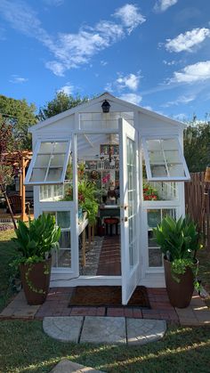 a small white greenhouse with potted plants in the front and side doors open on a sunny day