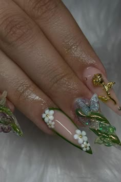 These stiletto nails transform into a miniature garden of enchantment. A beautiful nude base sets the stage for the creative 3D elements that spring to life. One nail is home to a delicate butterfly with translucent wings poised for flight, while metallic gold embellishments add a touch of luxury. Another nail features an intricate rose, its petals meticulously sculpted in gold, evoking a sense of timeless elegance. The floral theme continues with tiny white blossoms and pearl accents nestled among vibrant green leaves, giving a fresh and natural vibe to the overall design. This manicure masterfully combines artistry and nature for an ethereal and dimensional look. 🌿🌸🦋✨  // Photo Credit: Instagram @pinksugarnails_ Nails 3d Butterfly, Nails With Embellishments, Chrome Fairy Nails, Butterfly Garden Nails, Cottagecore Aesthetic Nails, Garden Inspired Nails, 3d Stiletto Nails, Floral Stiletto Nails, Nature Theme Nails