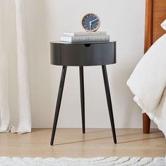 a small black table with a clock on it next to a white bed and curtains