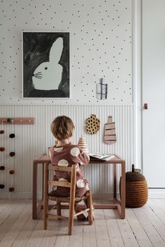 a little boy sitting at a table in front of a wallpapered room with a rabbit painting on the wall