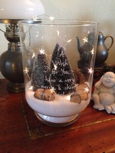 a glass filled with snow and trees on top of a wooden table next to a lamp