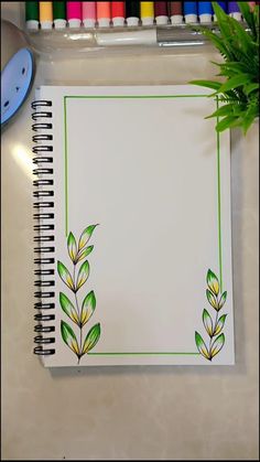 an open notebook with green leaves on it next to some crayon markers and a potted plant