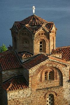 an old church with red tile roof next to the ocean and blue water in the background