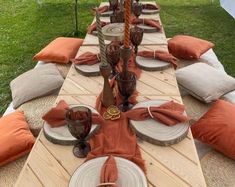 an outdoor table set up with place settings and orange napkins on it, in the middle of a grassy area