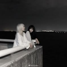 two women sitting on the edge of a pier at night with city lights in the background