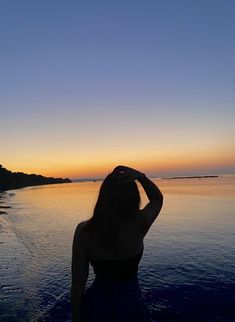 a woman standing on top of a beach next to the ocean at sunset with her hands in her hair