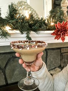 a person holding up a drink in front of a fireplace with christmas decorations on the mantle