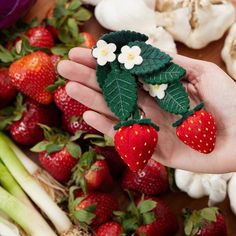 a hand holding a strawberry brooch with flowers and leaves on it next to vegetables