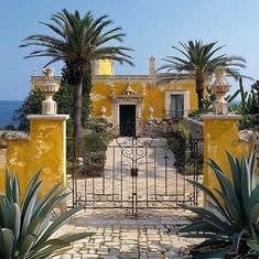 an iron gate leads to a yellow house with palm trees in the foreground and ocean in the background