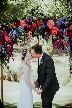 a man and woman are kissing under an arch with colorful flowers on it in front of them