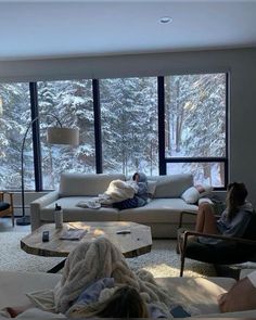 two people sitting on couches in a living room with snow covered trees outside the window