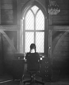 a woman sitting at a desk in front of a large window with a chandelier hanging from the ceiling