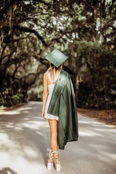 a woman wearing a green graduation cap and gown walks down the road in front of trees