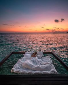 a woman laying on top of a bed in the middle of the ocean at sunset