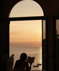 a woman sitting in front of an open window looking out at the ocean while reading a book