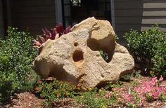 a large rock sitting in the middle of a flower garden next to a house with flowers