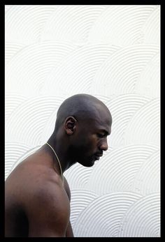 a man with no shirt on standing in front of a white wall and wearing a gold necklace