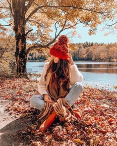 a woman sitting on the ground next to a tree with leaves all over her and wearing a red knitted hat