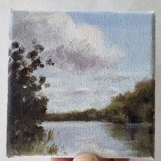 someone is holding up an oil painting of a lake in the middle of their hand