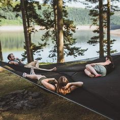 three people laying in a hammock on the grass near water and trees,