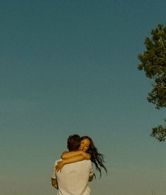 two people hugging each other in the middle of a field with a tree behind them