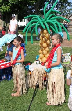 two girls in hula skirts are standing next to a palm tree and other people