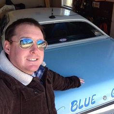 a man wearing sunglasses sitting in front of a blue car